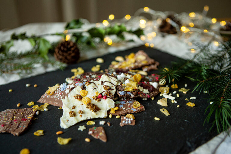 Op een plaat van leisteen ligt gebroken chocolade, op de achtergrond ziet men dennentakken en een lichtketting.