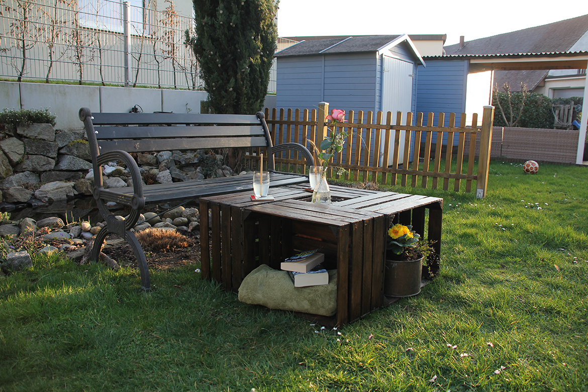 De tuintafel van houten kisten staat op het gazon voor een tuinbank.