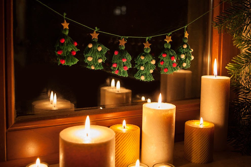 De slinger met kerstboompjes van garen hangt aan een raam met kaarsen op de vensterbank.